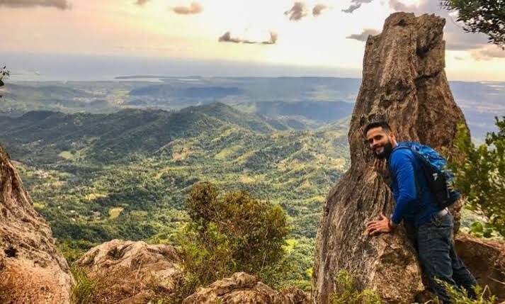 Un caminante con mochila se apoya en una roca alta frente a un valle verde y montañas al atardecer